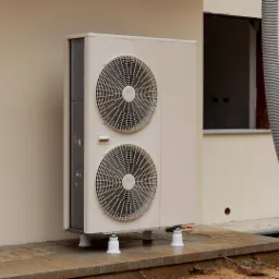 Zewnętrzna jednostka pompy ciepła, zamontowana na białych podstawkach na kostce brukowej w Miliczu, widoczna ściana budynku z oknem.