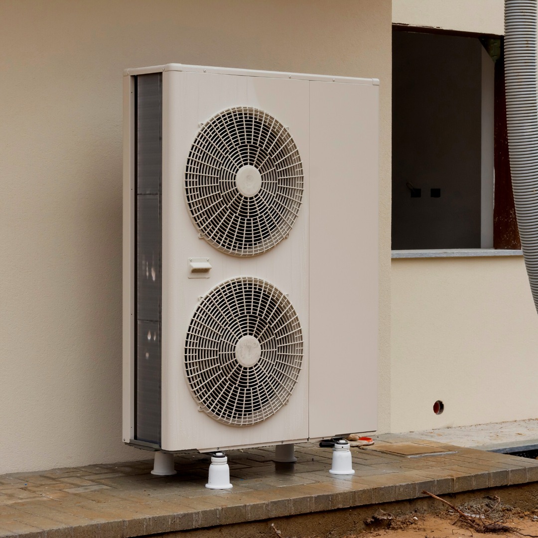 Zewnętrzna jednostka pompy ciepła, zamontowana na białych podstawkach na kostce brukowej w Miliczu, widoczna ściana budynku z oknem.