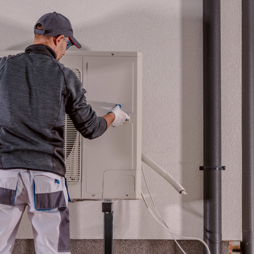 Mężczyzna w szarym ubraniu i czapce instaluje klimatyzator na ścianie o chropowatej fakturze, widoczne rury i izolacja.