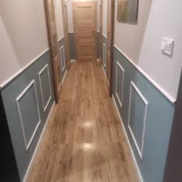 Korytarz po remoncie z panelami ściennymi w kolorze gołębim, drewnianą podłogą i drzwiami w naturalnym odcieniu drewna, zakończony obrazem z motywem leśnym.