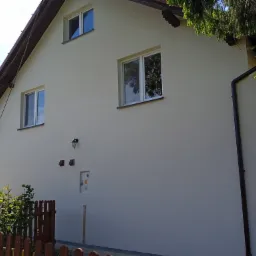 Jasna elewacja domu jednorodzinnego po tynkowaniu, widoczne okna z odbijającym się niebem, brązowe rynny i fragment ogrodzenia.