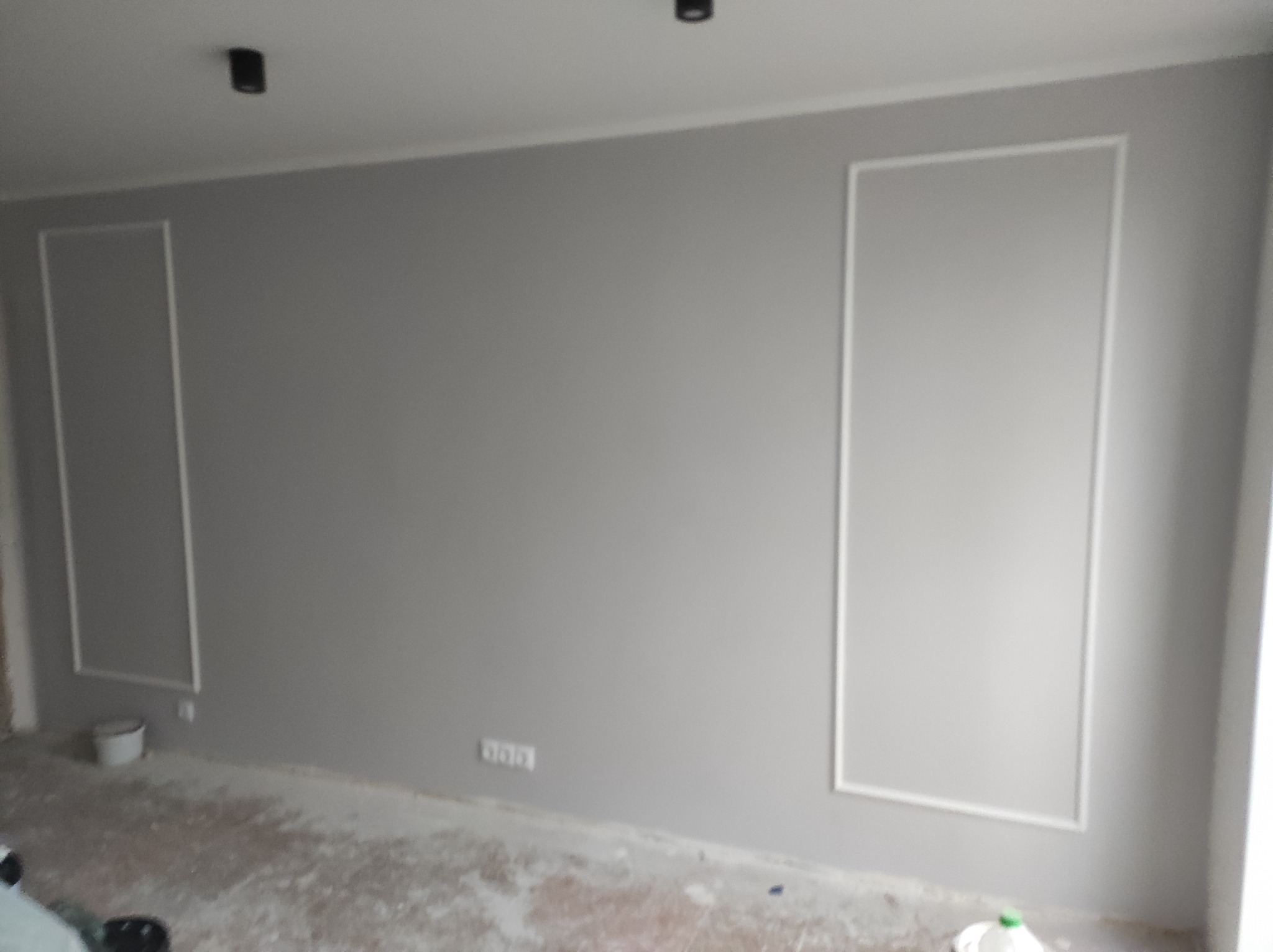 Szara ściana z białymi dekoracyjnymi ramkami, widoczne gniazdka elektryczne i ślady po remoncie na podłodze, sufit z dwoma czarnymi punktami oświetleniowymi.