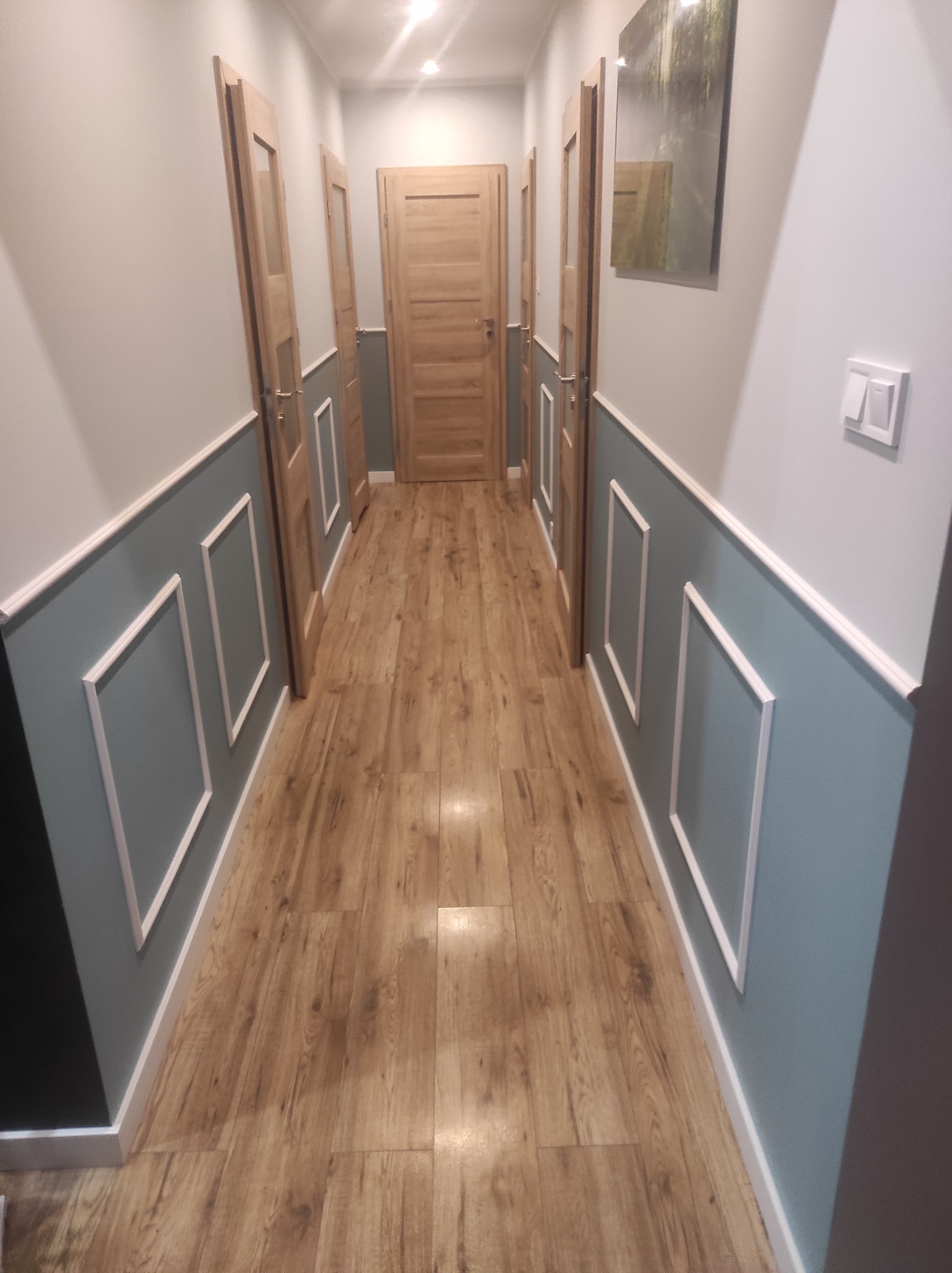 Korytarz po remoncie z panelami ściennymi w kolorze gołębim, drewnianą podłogą i drzwiami w naturalnym odcieniu drewna, zakończony obrazem z motywem leśnym.