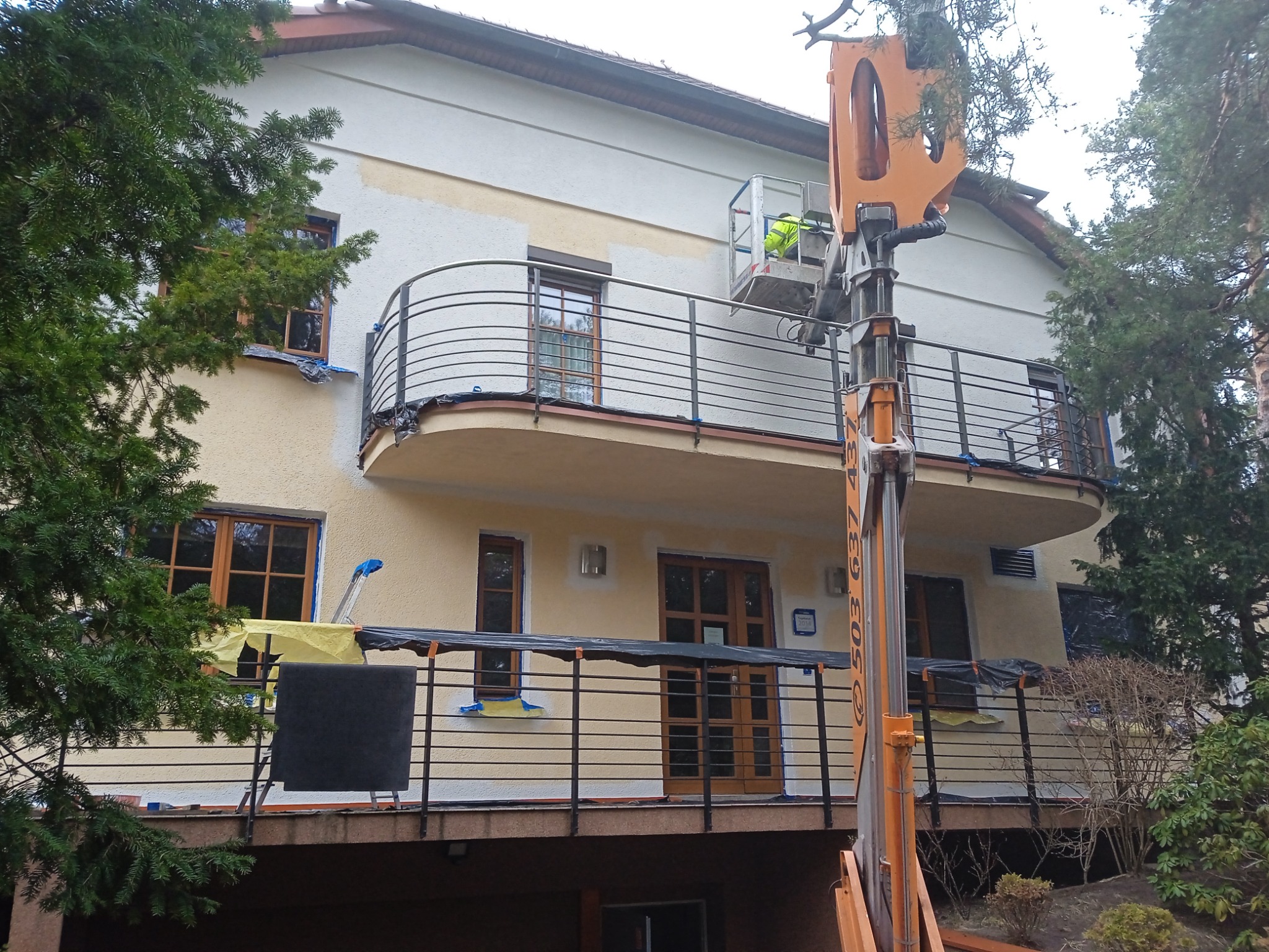 Dwupiętrowy budynek w trakcie odnawiania elewacji, widoczny podnośnik koszowy z pracownikiem, fragmenty pomalowanej i niepomalowanej ściany, zabezpieczone okna i balkon.