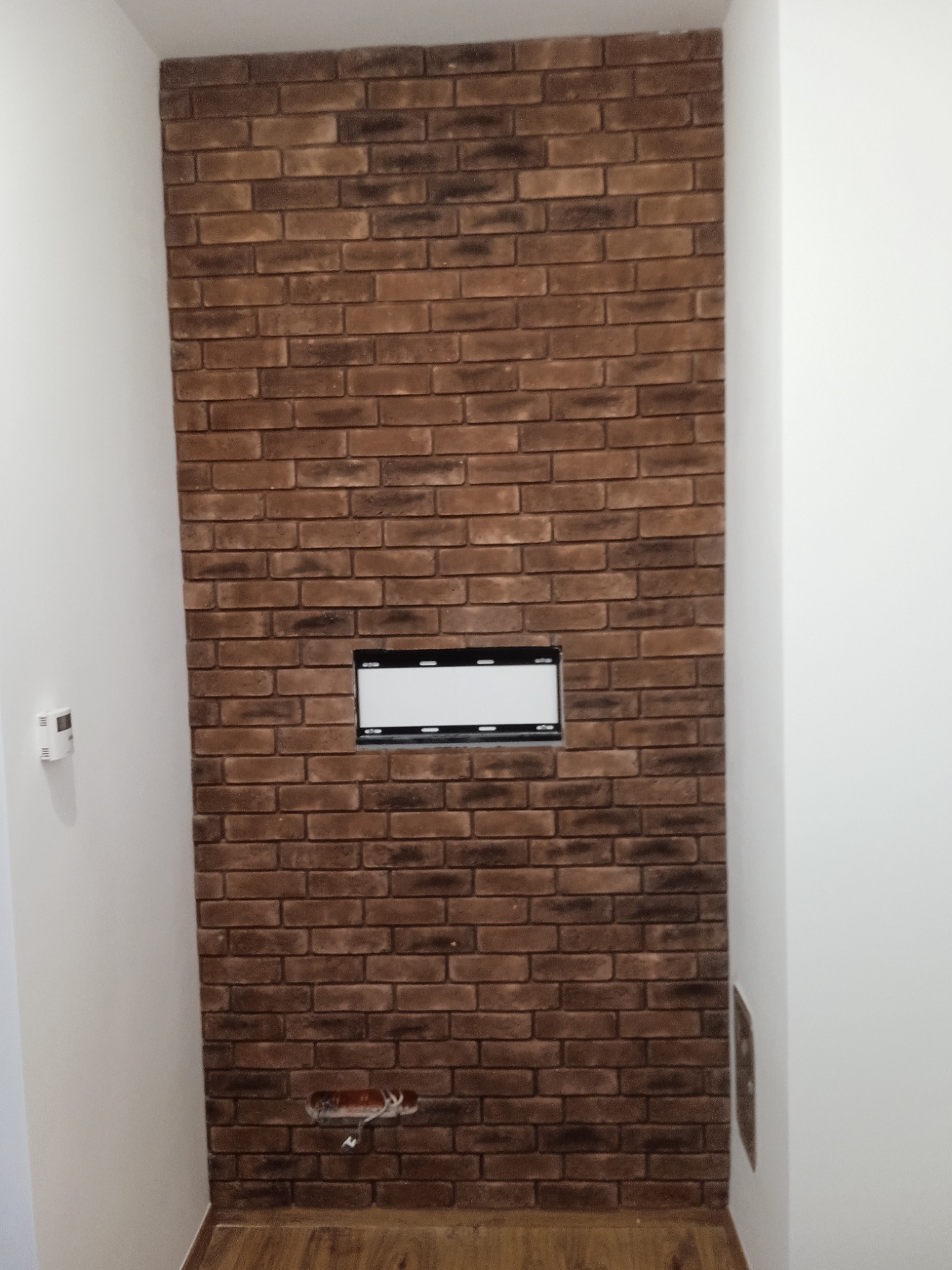 Ściana wykończona cegłą dekoracyjną w odcieniach brązu, z widocznym otworem na instalację elektryczną i puszką pod gniazdko, otoczona białymi ścianami.