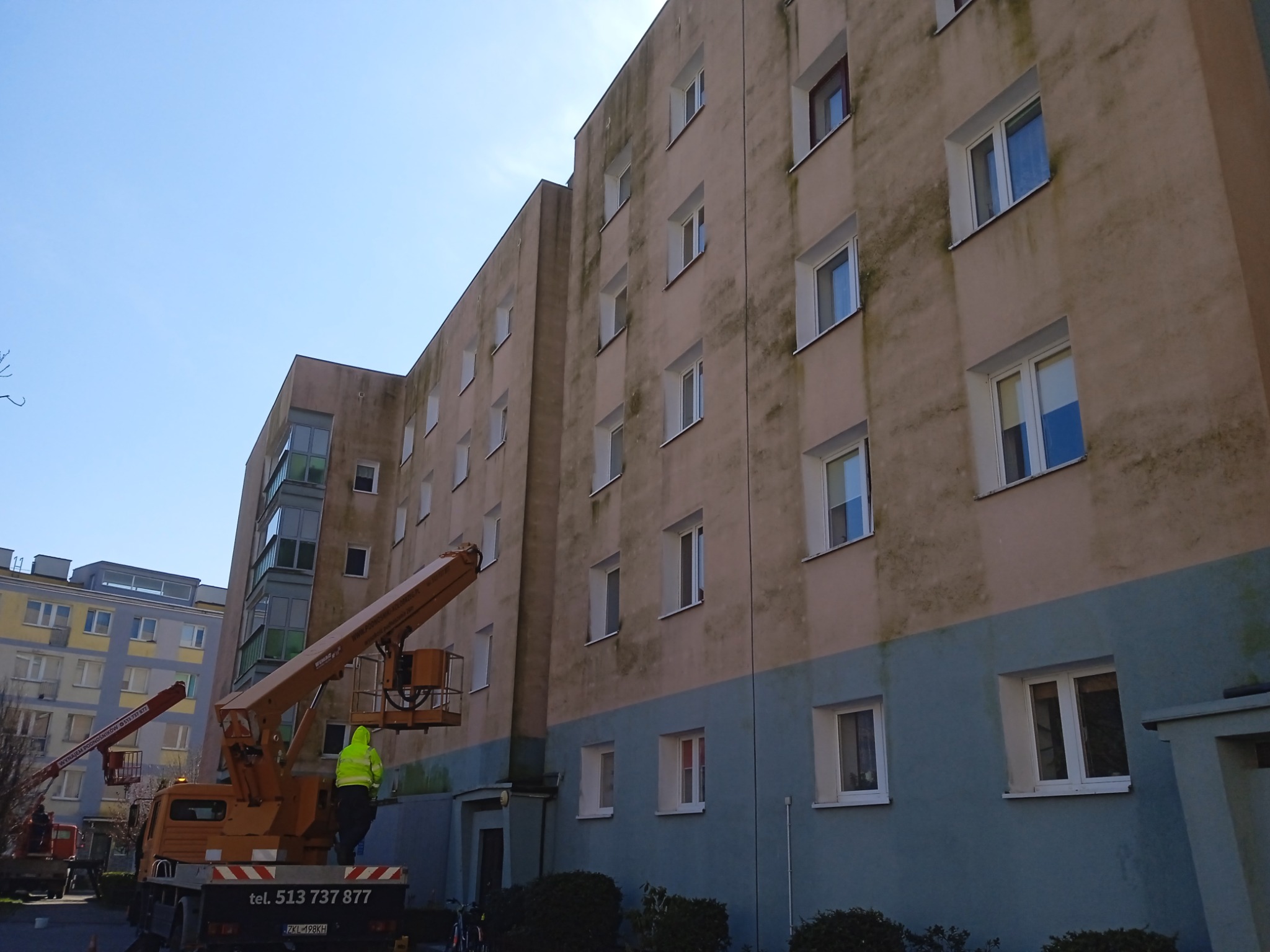 Pracownik w żółtej kurtce obsługuje podnośnik koszowy podczas renowacji elewacji bloku mieszkalnego, widoczne ślady zabrudzeń i różnica kolorów na ścianie.