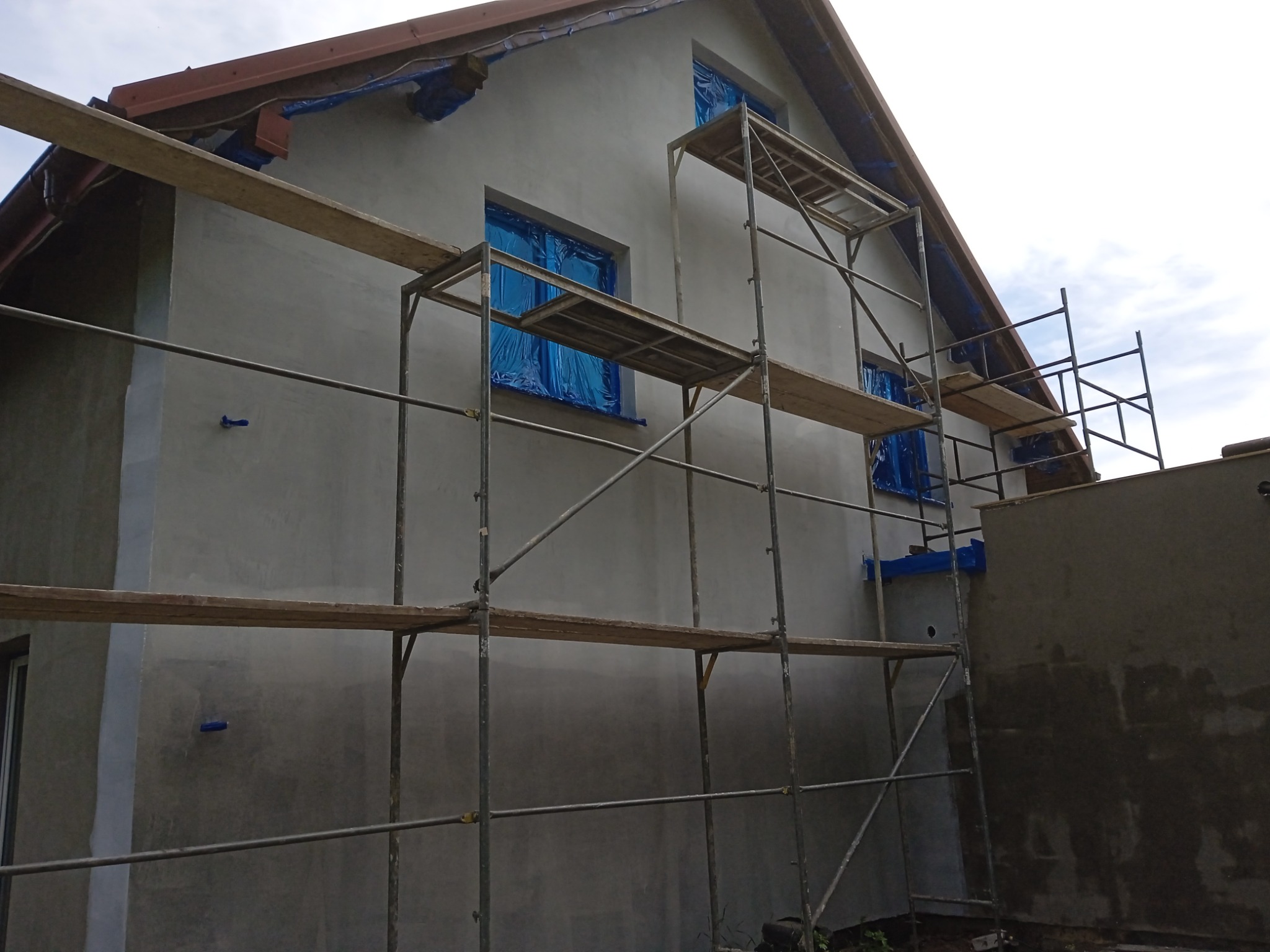 Dom w trakcie tynkowania elewacji, okna zabezpieczone folią, rusztowanie metalowe, widoczne ślady tynku na ścianie.