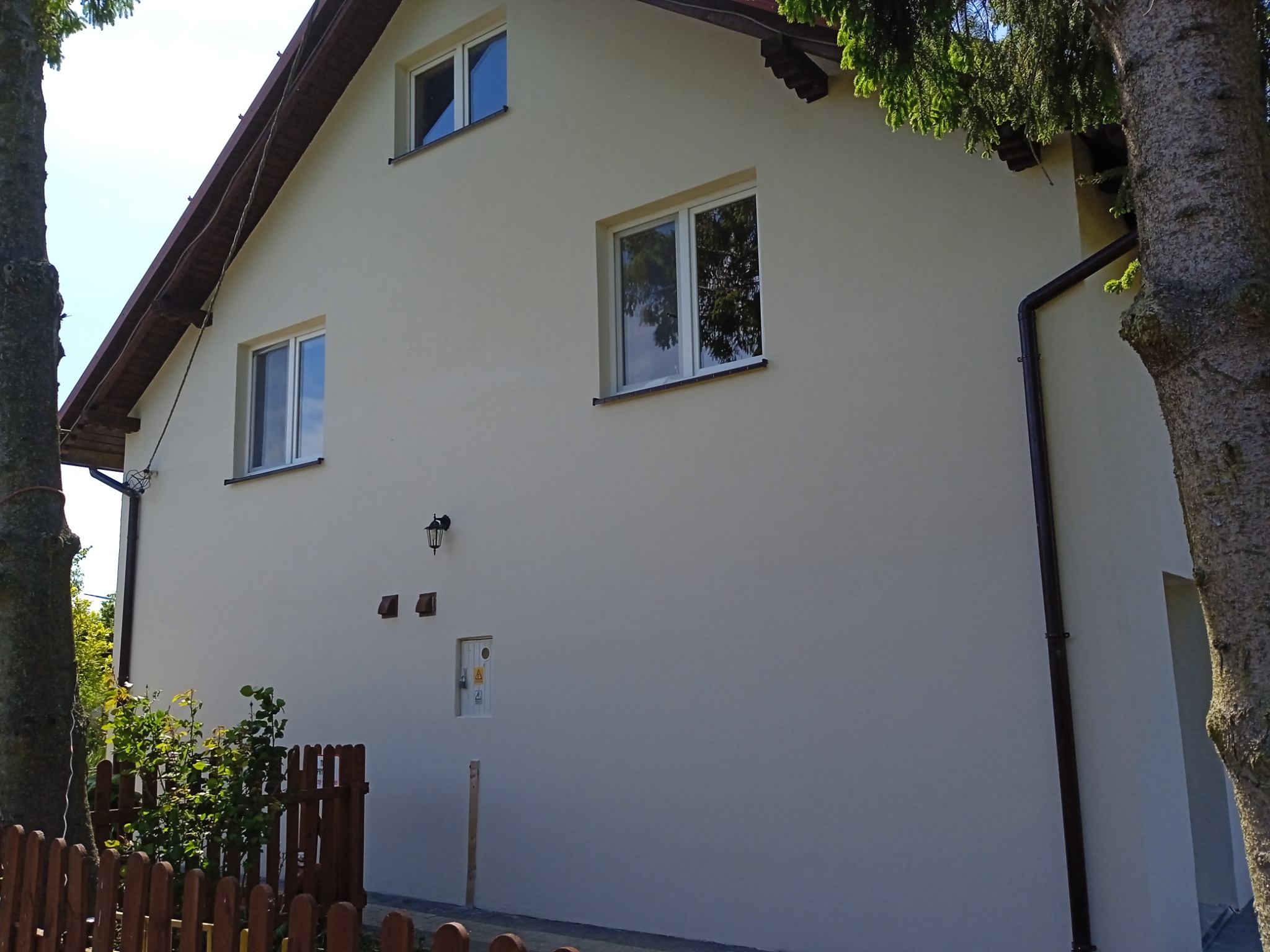 Jasna elewacja domu jednorodzinnego po tynkowaniu, widoczne okna z odbijającym się niebem, brązowe rynny i fragment ogrodzenia.