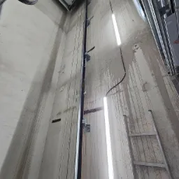 Wnętrze szybu windowego w trakcie montażu, widoczne metalowe prowadnice, oświetlenie tymczasowe w postaci lamp liniowych oraz drabina oparta o surową ścianę.