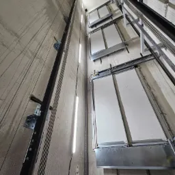 Wnętrze szybu windy w trakcie montażu: widoczne szyny prowadzące, oświetlenie tymczasowe oraz mechanizm drzwi szybowych z białymi panelami.