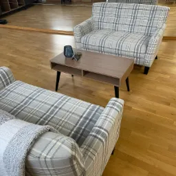 Dwa fotele i stolik kawowy w pokoju z parkietem i lustrem na ścianie. Fotele tapicerowane w szarą kratę, na jednym leży pled w zygzaki. Na stoliku wazon i wizytówki.