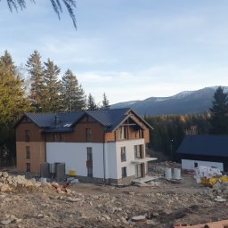 HUBELL SPÓŁKA Z OGRANICZONĄ ODPOWIEDZIALNOŚCIĄ - Dwukondygnacyjny dom z ciemnym dachem w trakcie budowy, otoczony gruzem i ziemią. W tle las iglasty i góry ze śniegiem. Widok z dołu, wczesne popołudnie.