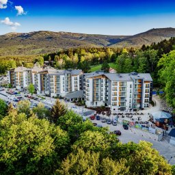 HUBELL SPÓŁKA Z OGRANICZONĄ ODPOWIEDZIALNOŚCIĄ - Kompleks apartamentów z szarymi dachami, widok z góry na tle zalesionych gór i błękitnego nieba. Parking przed budynkami.