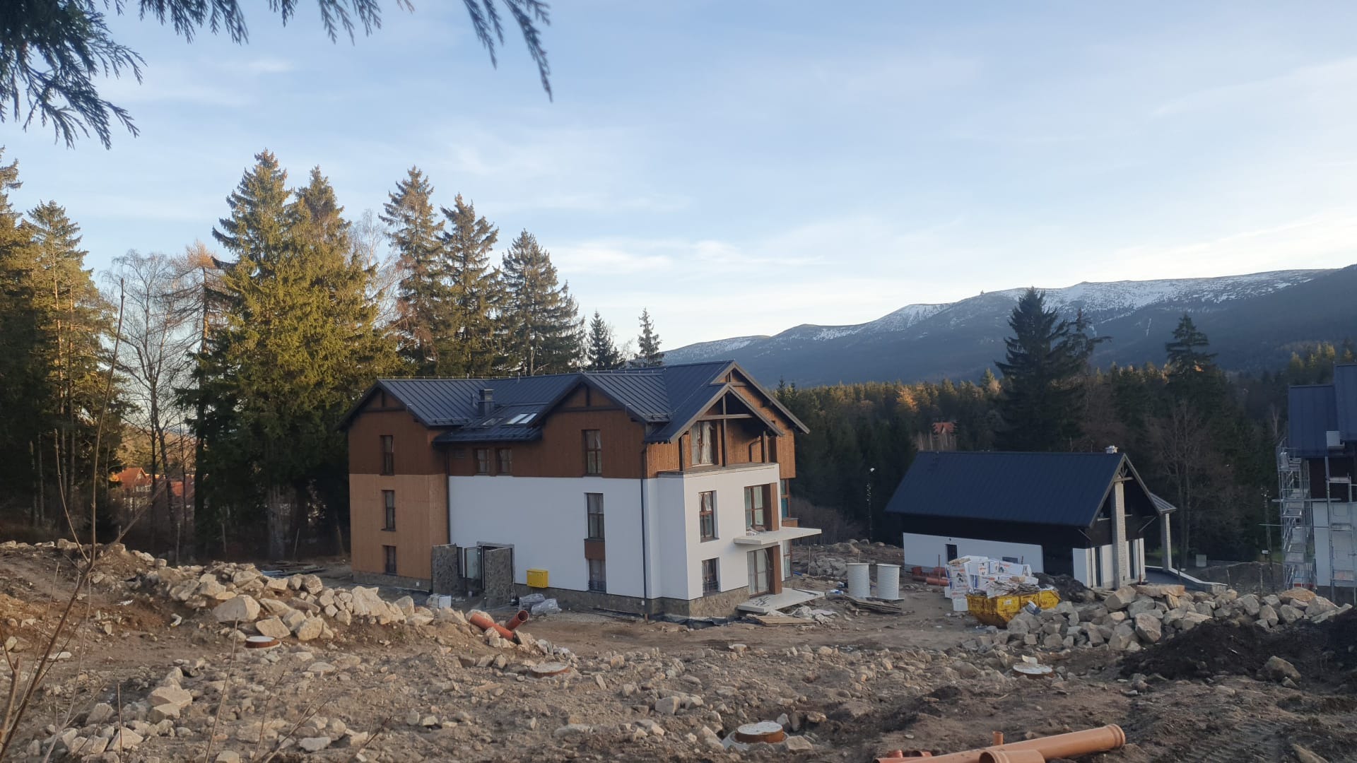 Dwukondygnacyjny dom z ciemnym dachem w trakcie budowy, otoczony gruzem i ziemią. W tle las iglasty i góry ze śniegiem. Widok z dołu, wczesne popołudnie.