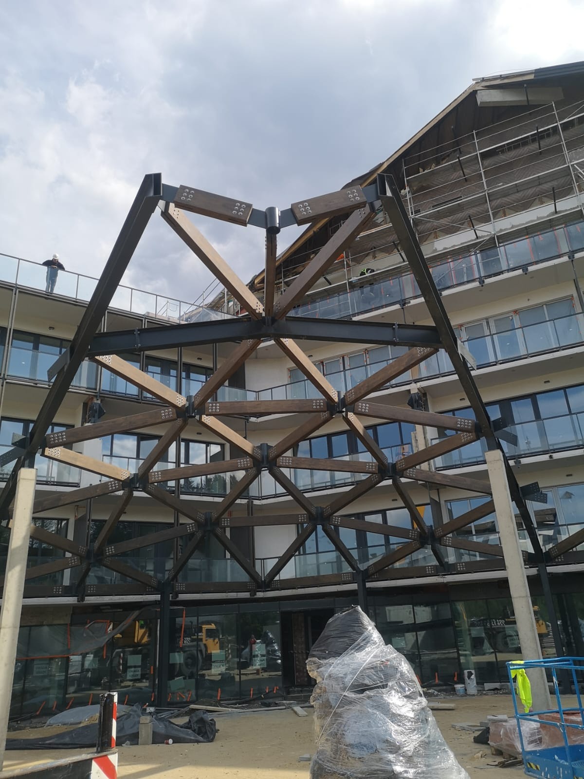 Konstrukcja dachu z drewna i stali na tle budynku z balkonami, widok z dołu. Elementy konstrukcyjne połączone śrubami. Na dachu rusztowania.