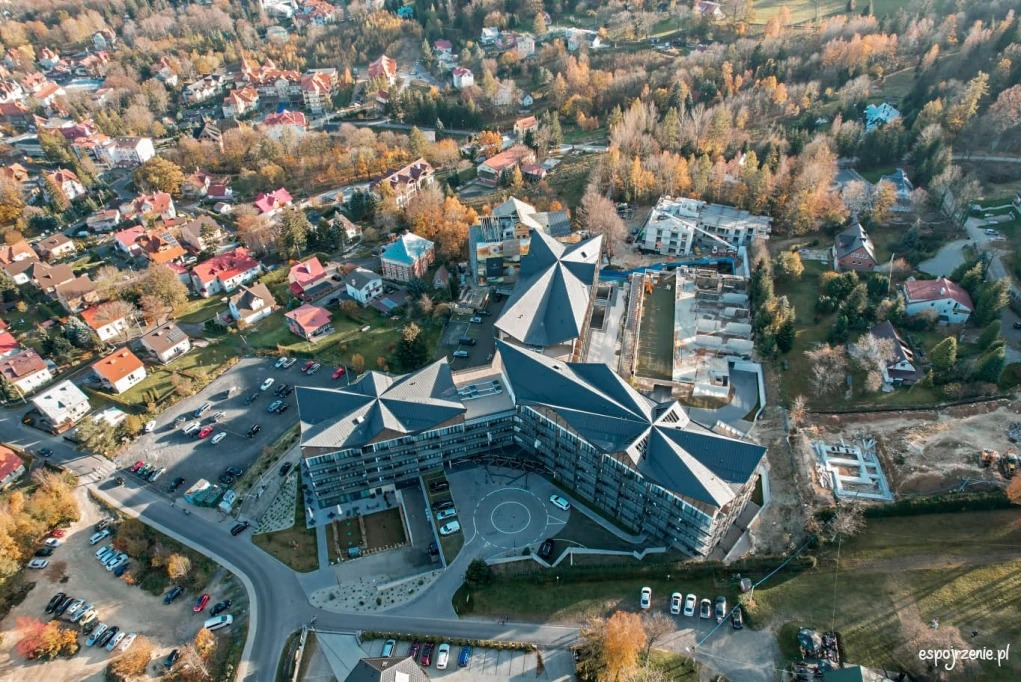 Widok z lotu ptaka na duży budynek z ciemnymi dachami o geometrycznym kształcie, otoczony drzewami i zabudową mieszkalną w jesiennej scenerii.