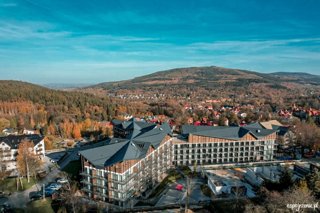 Nowoczesny budynek apartamentowy z ciemnym dachem na tle górskiego krajobrazu w jesiennej scenerii. Widok z lotu ptaka, błękitne niebo.