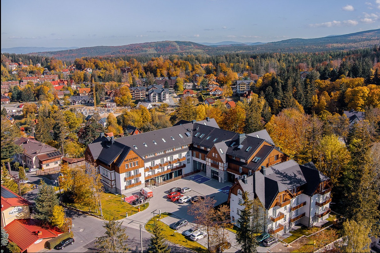 Widok z lotu ptaka na hotel z ciemnym dachem w otoczeniu jesiennych drzew i zabudowy miejskiej. W tle góry i błękitne niebo.