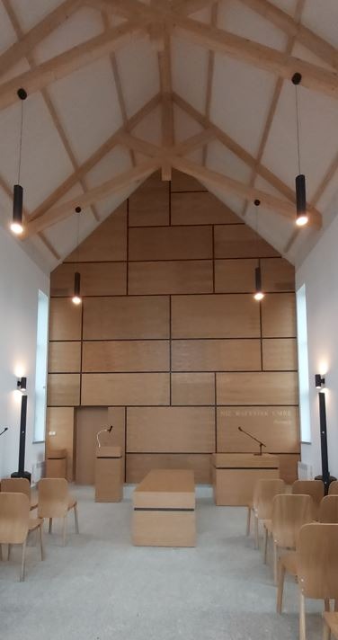 Jasne wnętrze z drewnianą konstrukcją dachu i panelami ściennymi. Widoczne krzesła i mównice, oświetlenie punktowe. Minimalistyczny design.