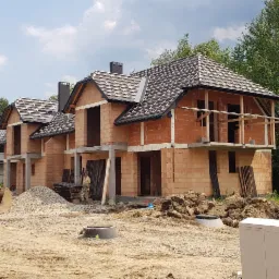 Surowe domy w trakcie budowy z cegły ceramicznej z widocznym szkieletem dachu i elementami konstrukcyjnymi balkonu, na pierwszym planie plac budowy z hałdą ziemi i materiał&oacute;w budowlanych.