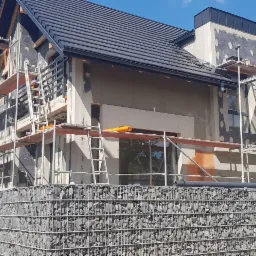 Dom jednorodzinny w trakcie prac elewacyjnych, widoczne rusztowania, ocieplenie oraz kamienne umocnienie skarpy.