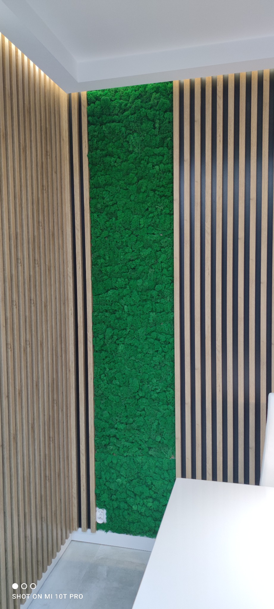 Ściana z panelami drewnianymi i panelem z mchu dekoracyjnego w kolorze intensywnej zieleni, widoczne gniazdko elektryczne.