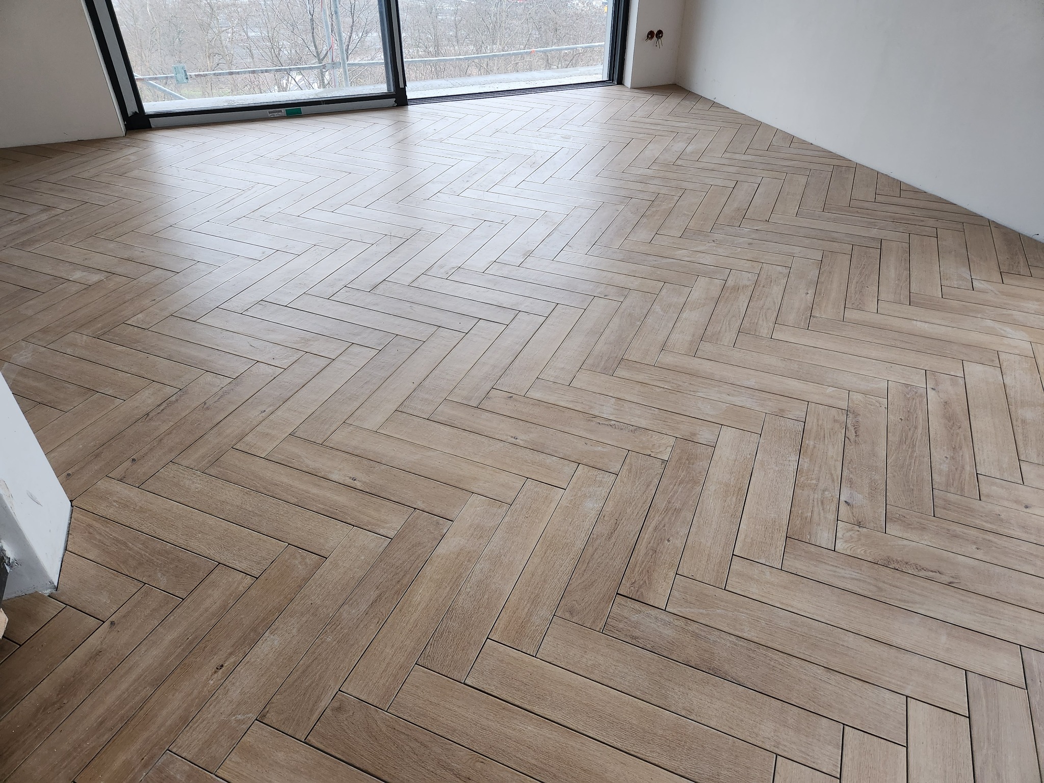 Ułożona parkietem jodełkowym podłoga z paneli w jasnobrązowym odcieniu drewna w pokoju z dużym oknem i surowymi ścianami.