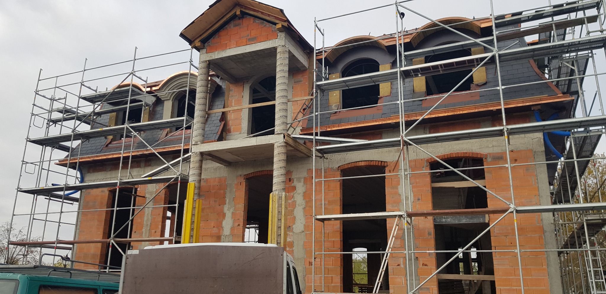 Budowa domu z cegły z elementami elewacji z łupka, widoczne rusztowania i przygotowane otwory okienne.
