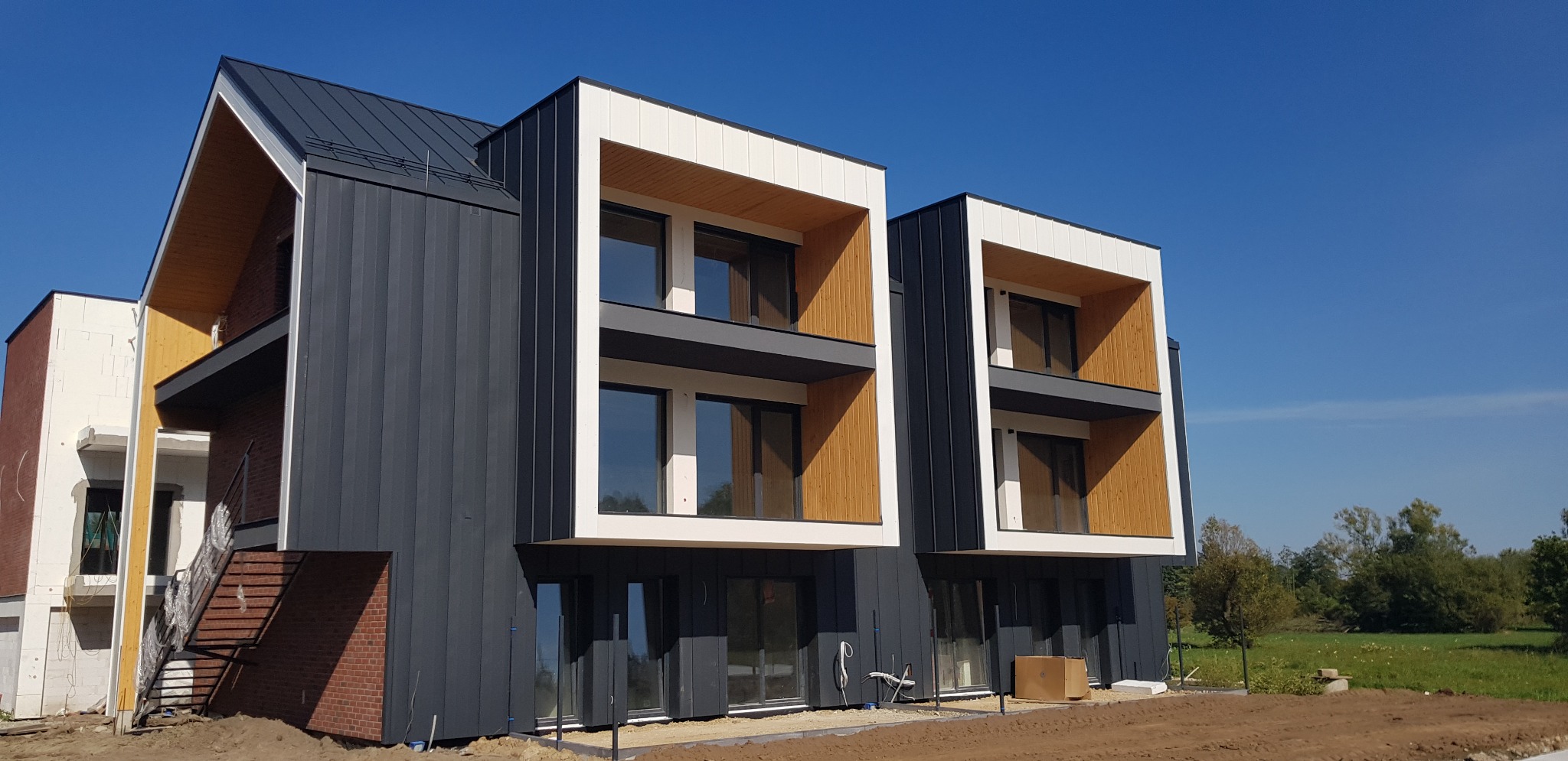 Nowoczesny budynek wielorodzinny w Krakowie z elewacją z paneli w kolorze grafitowym, z białymi i drewnianymi akcentami w wykończeniu okien i balkonów, widoczny na tle błękitnego nieba i zieleni.