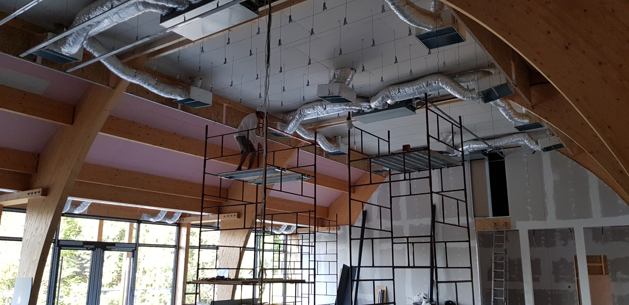 Montaż sufitu podwieszanego w dużej hali z widocznymi elementami wentylacji, pracownik na rusztowaniu, konstrukcja drewniana dachu.