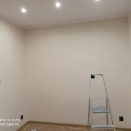 Świeżo pomalowane wnętrze pokoju w odcieniach beżu, widoczne gniazdka elektryczne, aluminiowa drabina i fragment grzejnika z żółtą szmatką.