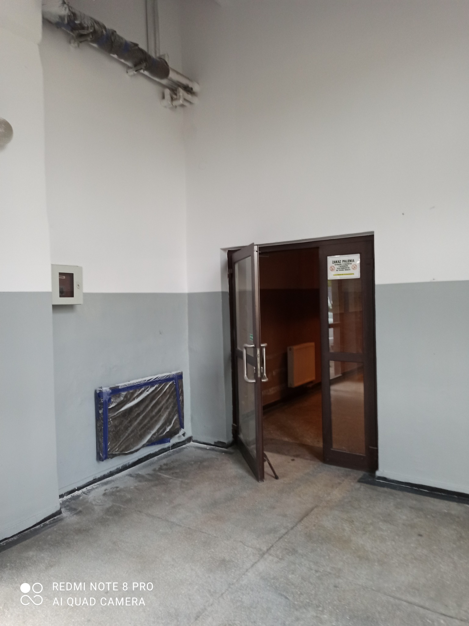 Przygotowanie do malowania: Zakryty grzejnik folią malarską w pomieszczeniu z dwukolorowymi ścianami i otwartymi drzwiami.