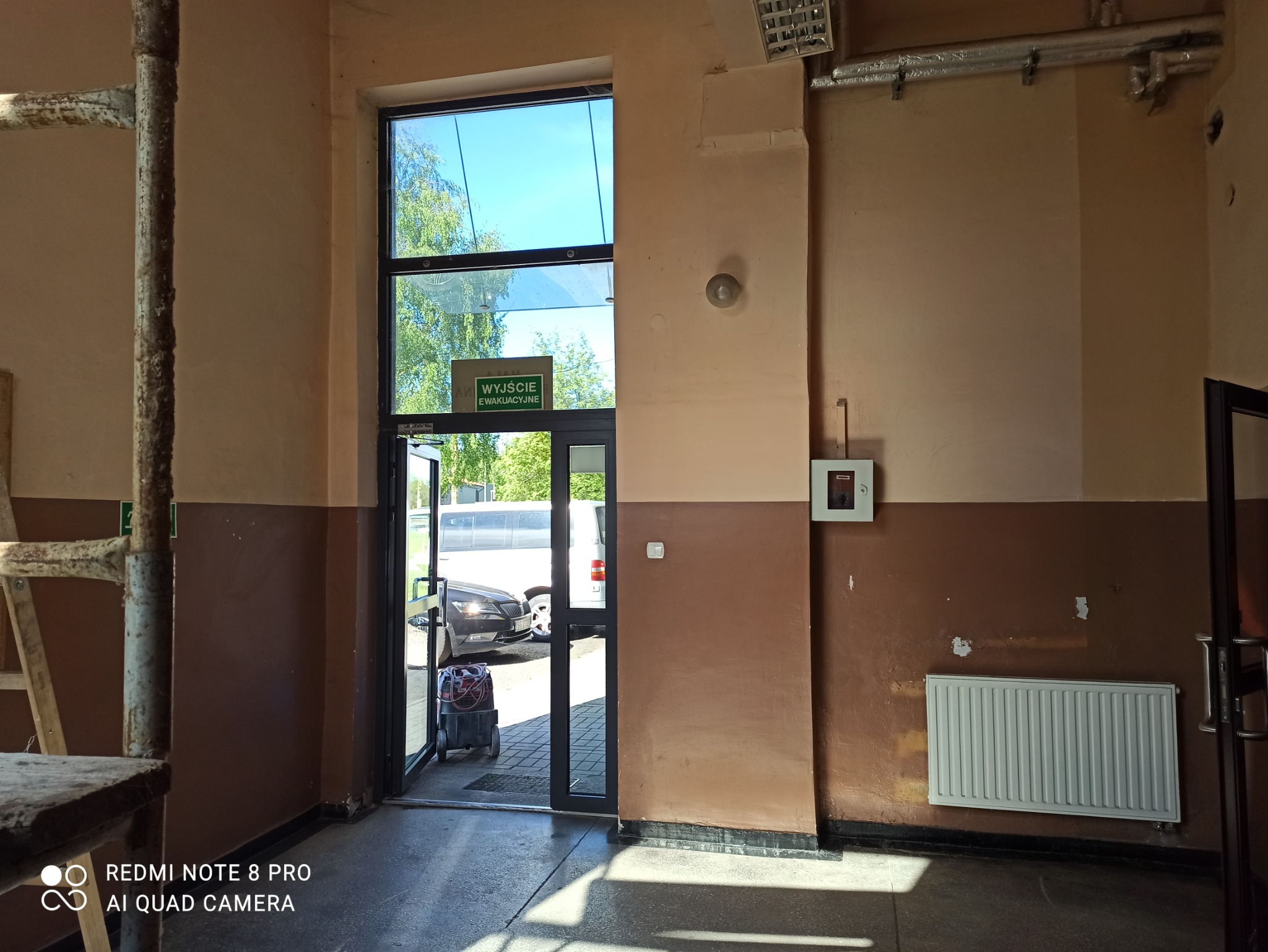 Wnętrze budynku w trakcie remontu, widoczne ślady po naprawach na ścianach, grzejnik, otwarte drzwi ewakuacyjne z widokiem na zewnątrz, drabina malarska i odkurzacz przemysłowy.