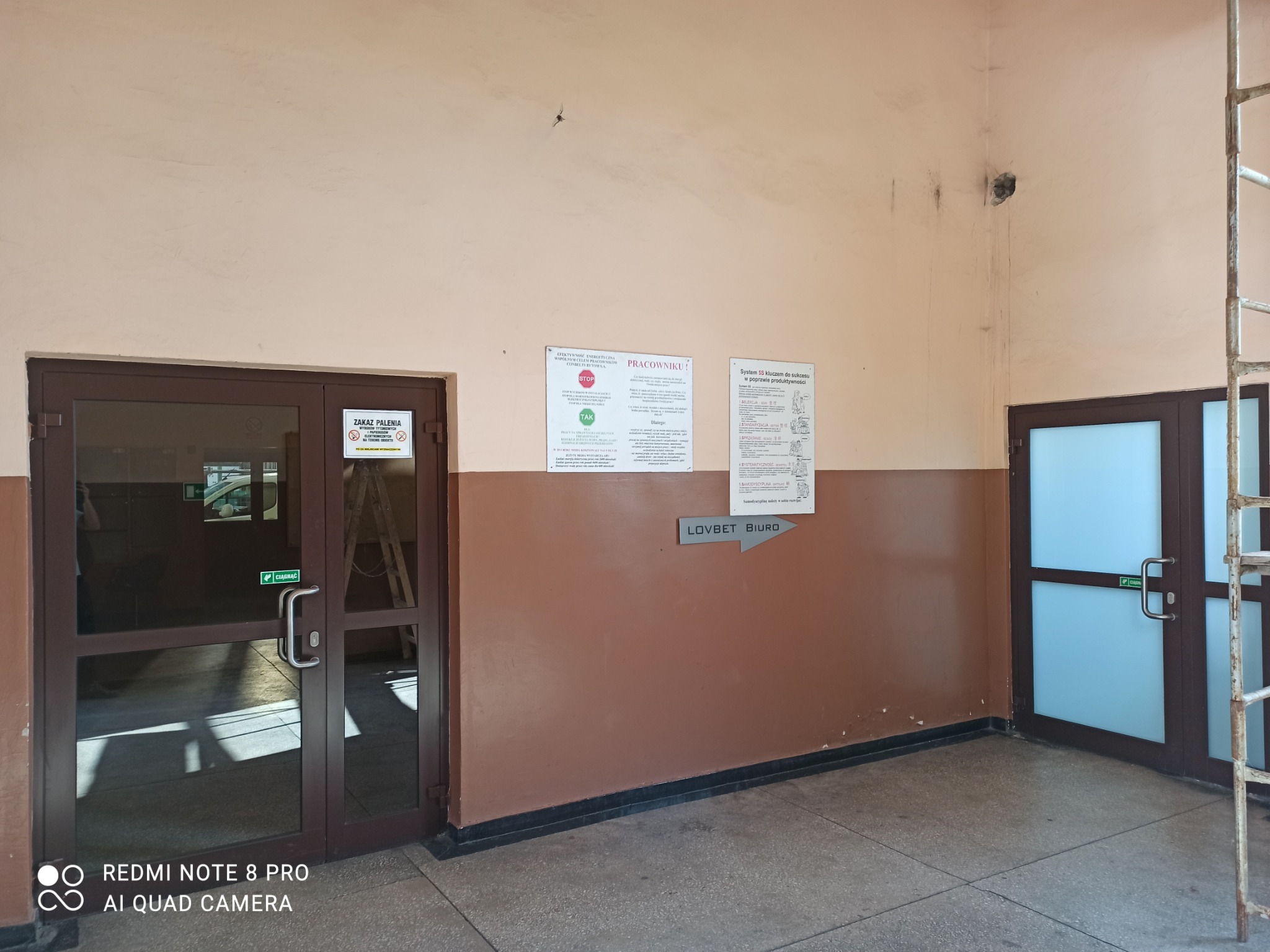 Wnętrze biura z dwoma parami szklanych drzwi, tablicami informacyjnymi na ścianie i drabina oparta o ścianę.