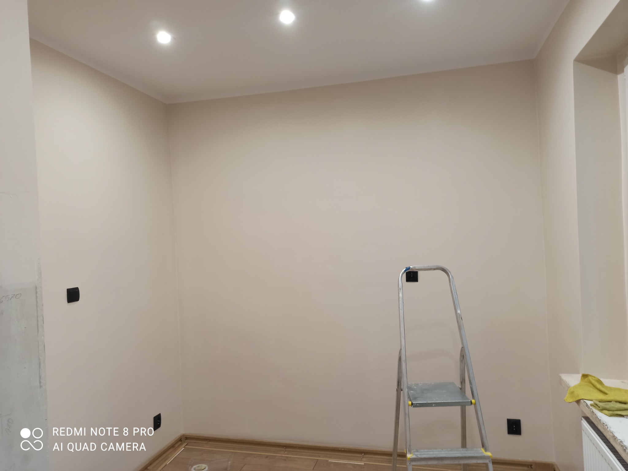 Świeżo pomalowane wnętrze pokoju w odcieniach beżu, widoczne gniazdka elektryczne, aluminiowa drabina i fragment grzejnika z żółtą szmatką.