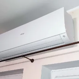 Biały klimatyzator Haier zamontowany na ścianie nad oknem, z widocznym karniszem z ciemnego metalu z żabkami.