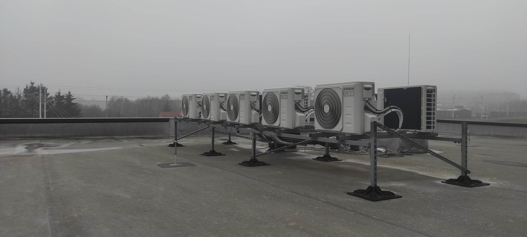 Rząd jednostek zewnętrznych klimatyzacji Kaisai Eco, ustawionych na metalowej konstrukcji na dachu budynku w pochmurny dzień.
