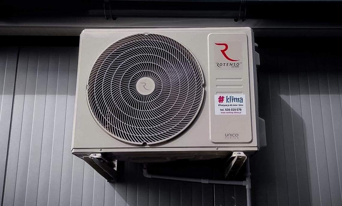 Zewnętrzna jednostka klimatyzacji Rotenso Unico Series zamontowana na szarej, metalowej ścianie z widocznym hashtagiem #klima i numerem telefonu.