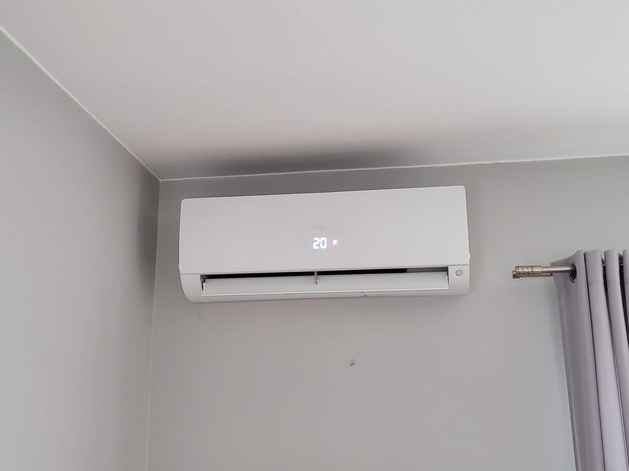 Biały klimatyzator ścienny z wyświetlaczem temperatury 20 stopni Celsjusza, zamontowany w szarym pokoju, widoczne fragmenty zasłon.