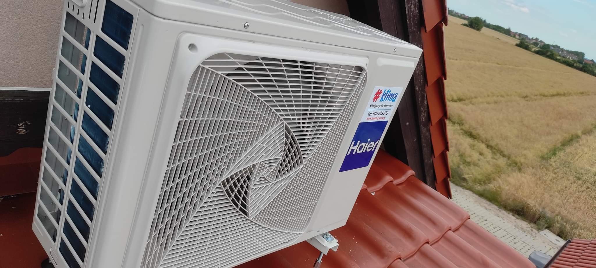 Zewnętrzna jednostka klimatyzacji Haier zamontowana na czerwonym dachu domu jednorodzinnego z widokiem na pole.