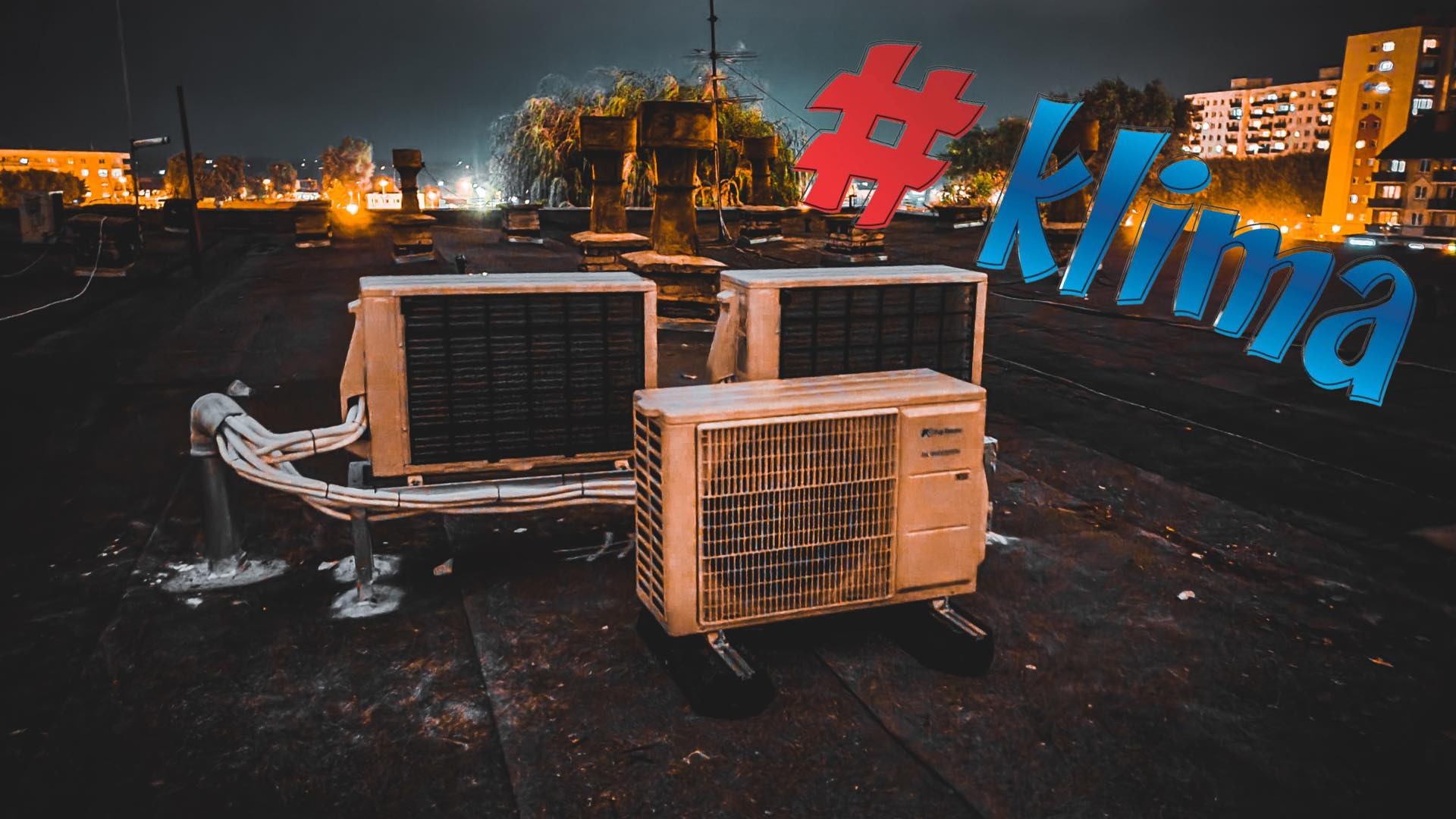 Trzy zewnętrzne jednostki klimatyzacyjne na dachu budynku w nocnej scenerii miejskiej, z widocznymi przewodami i elementami instalacji oraz nałożonym tekstem '#Klima'.