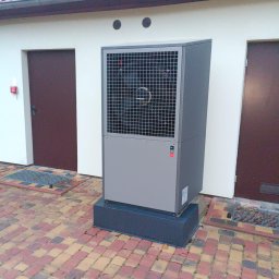 Instal-SUN Sąsiedzka Energia - Szara pompa ciepła powietrze-woda na zewnątrz budynku z czerwonym dachem i dwoma brązowymi drzwiami. Kostka brukowa w odcieniach czerwieni i beżu. Kratka odpływowa.