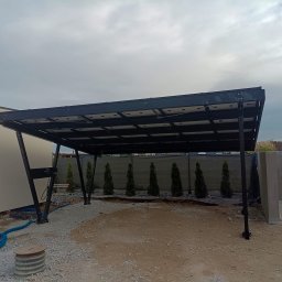 Instal-SUN Sąsiedzka Energia - Zadaszenie z paneli, prawdopodobnie fotowoltaicznych, na metalowej konstrukcji. Widoczne kable i rury, sugerujące podłączenie do instalacji. W tle rząd małych drzewek.