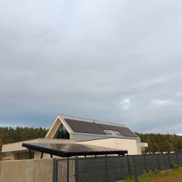 Instal-SUN Sąsiedzka Energia - Nowoczesny dom jednorodzinny z panelami fotowoltaicznymi na dachu i wiatą solarną w Rawiczu. Widok zza ogrodzenia, pochmurne niebo w tle.