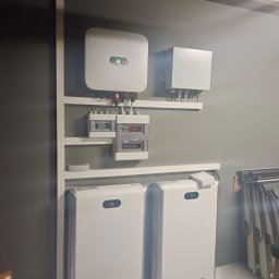 Instal-SUN Sąsiedzka Energia - Kompletna instalacja fotowoltaiczna w garażu: falownik, magazyn energii (dwie sztuki) i rozdzielnia elektryczna na ścianie. Widoczne okablowanie i elementy montażowe.