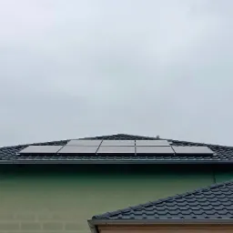 Część dachu pokryta czarną dachówką z zainstalowanymi panelami słonecznymi w układzie 2x3, widoczne orynnowanie i elewacja budynku w odcieniach zieleni, pochmurne niebo w tle.