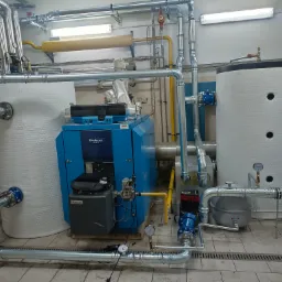 Kompleksowa instalacja kotłowni z niebieskim kotłem Buderus, dwoma białymi zbiornikami akumulacyjnymi oraz rozbudowanym systemem rur w pomieszczeniu technicznym z kratką ściekową w podłodze.