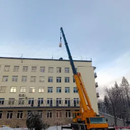 Żółty dźwig samochodowy podnosi ładunek na dach budynku w zimowy dzień, widoczne zasypane śniegiem drzewa i parking.