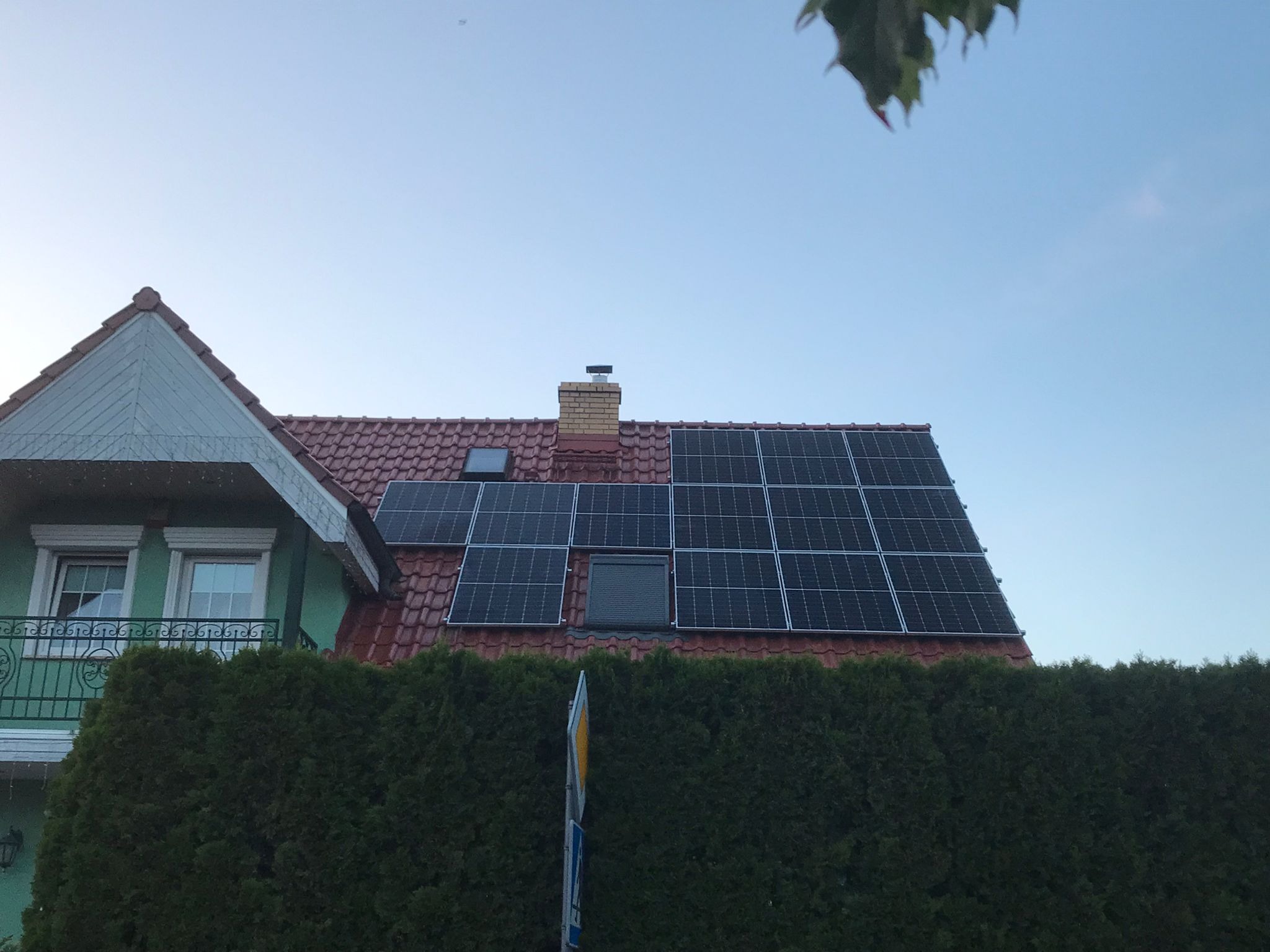 Panele słoneczne na dachu domu z czerwoną dachówką, widoczne okna dachowe i komin, na pierwszym planie żywopłot i znak drogowy.