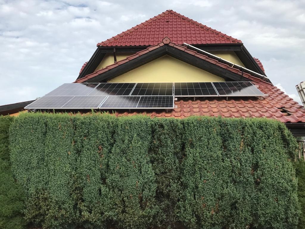 Panele słoneczne zamontowane na dachu domu z czerwoną dachówką, częściowo zasłonięte wysokim żywopłotem; widoczna drabina z boku.